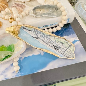 May include: A gold-painted oyster shell with a map of Seagrove Beach, Florida inside. The map shows the location of Watercolor, Seaside, and Canal Street. The oyster shell is sitting on a book with a white beaded necklace and other oyster shells.