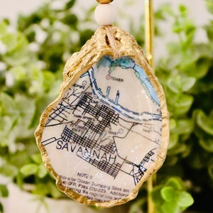 May include: A gold-painted oyster shell ornament with a map of Savannah, Georgia printed on it. The ornament is hanging from a gold string with white and gold beads.