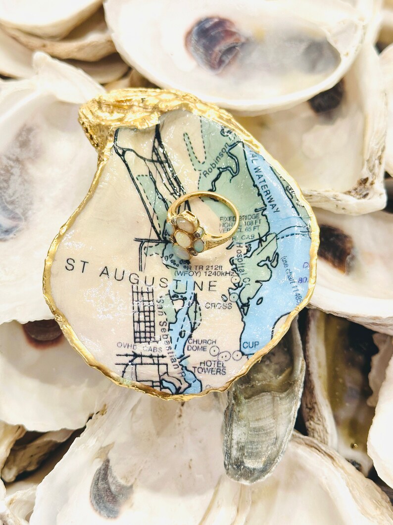 May include: A gold ring with opal stones sits on a decorative oyster shell. The shell is painted with a map of St. Augustine, Florida, and has a gold-painted edge. The ring dish is surrounded by other oyster shells.