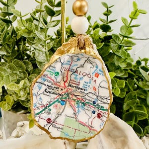 May include: An oyster shell ornament featuring a map of Tuscaloosa, Alabama. The shell is trimmed with gold paint and has a string for hanging, with wooden beads. The map includes roads and city names.