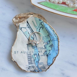 May include: An oyster shell ring dish with a map of St. Augustine, Florida, featuring blue and white details and gold trim. A silver ring rests inside the dish. A decorative plate and a string of pearls are in the background.