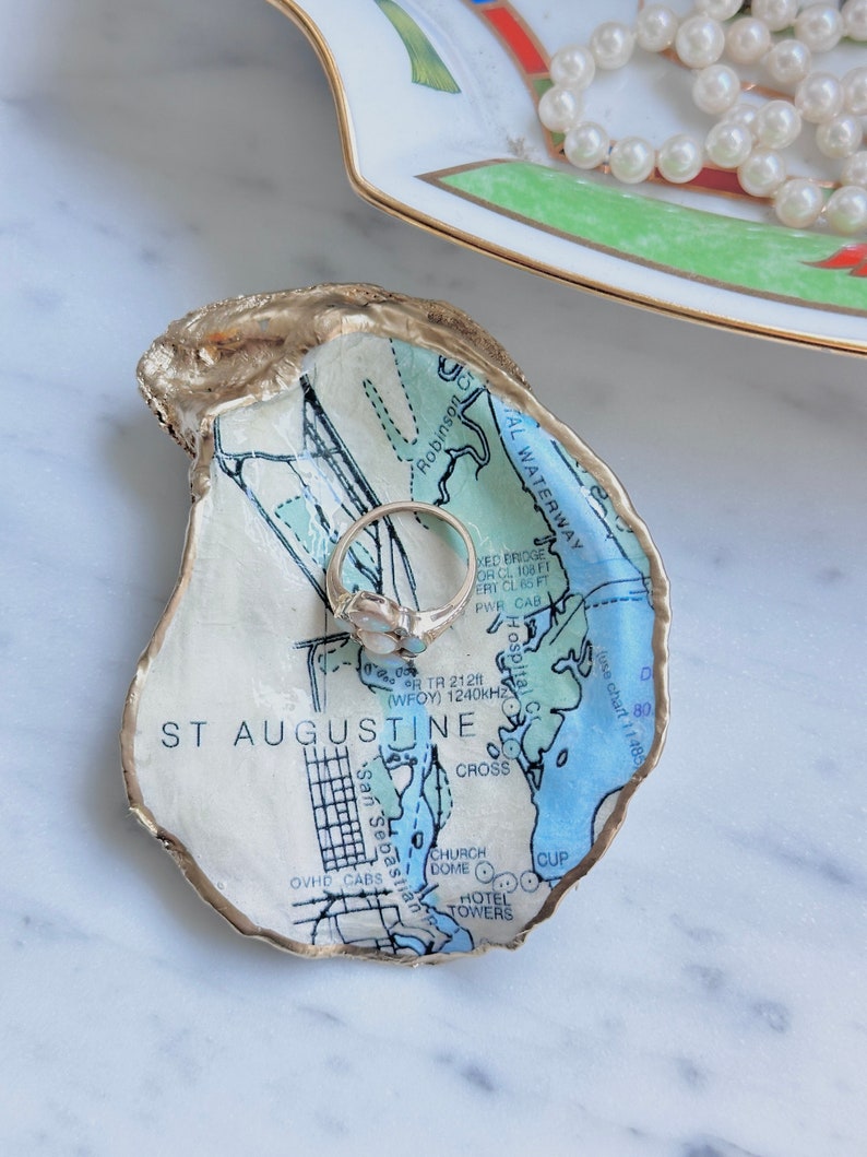 May include: An oyster shell ring dish with a map of St. Augustine, Florida, featuring blue and white details and gold trim. A silver ring rests inside the dish. A decorative plate and a string of pearls are in the background.