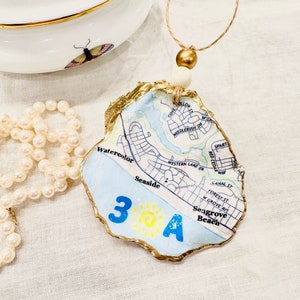 May include: A gold-painted oyster shell ornament with a map of Seagrove Beach, Florida. The ornament features the text "3A" and a sun graphic. The ornament is hanging from a twine cord with wooden beads.
