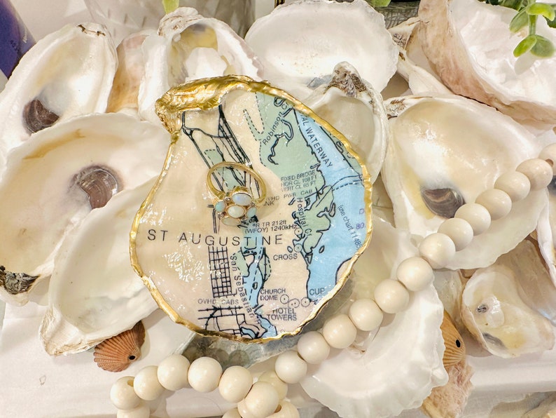 May include: An oyster shell ring dish featuring a map of St. Augustine, Florida, and a gold ring with white gemstones. The dish has a gold edge and is surrounded by other oyster shells and a beaded garland.