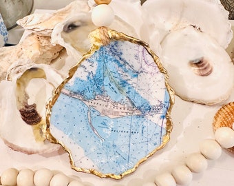May include: A gold-rimmed oyster shell ornament featuring a nautical map design. The ornament is suspended from a twine cord with wooden beads. The ornament is surrounded by white beads and oyster shells.