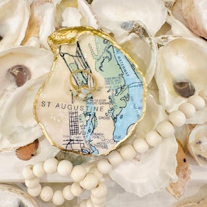 May include: An oyster shell ring dish with a gold-painted rim, featuring a map design of St. Augustine, Florida. A gold ring with white stones rests inside. The dish is surrounded by other oyster shells and a strand of white wooden beads.