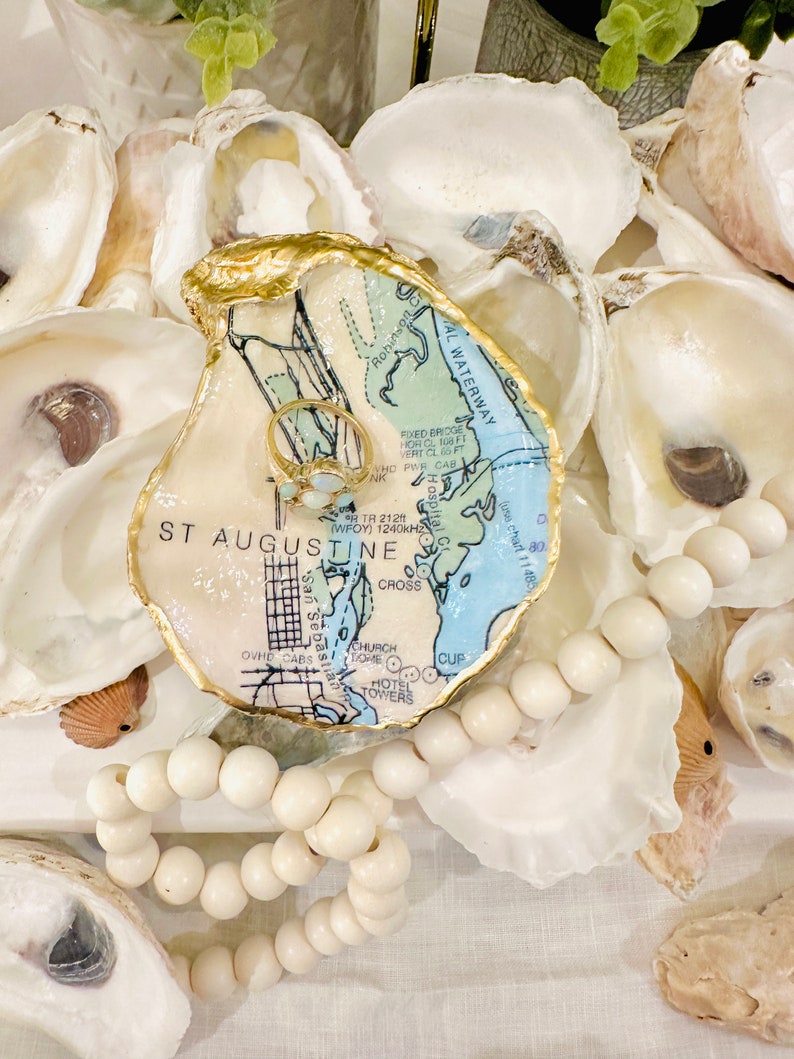 May include: An oyster shell ring dish with a gold-painted rim, featuring a map design of St. Augustine, Florida. A gold ring with white stones rests inside. The dish is surrounded by other oyster shells and a strand of white wooden beads.