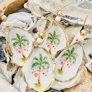 May include: Three oyster shell ornaments with gold trim, each featuring a painted palm tree with colorful ornaments hanging from the branches. The ornaments are strung with twine and wooden beads.