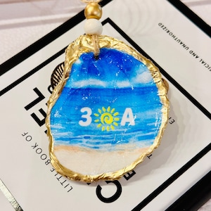 May include: A gold-painted seashell ornament with a blue and white painted seascape and the text "3 A" with a yellow sun in between the numbers.