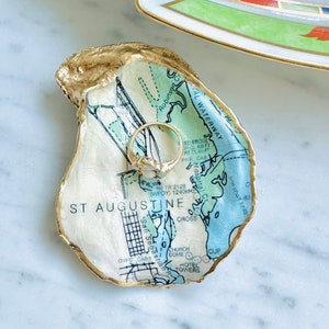 May include: An oyster shell ring dish featuring a map of St. Augustine, Florida. The shell has a gold-painted edge and holds a gold ring. The map design uses blue and green tones with black text. A decorative dish with a pearl necklace is in the background.