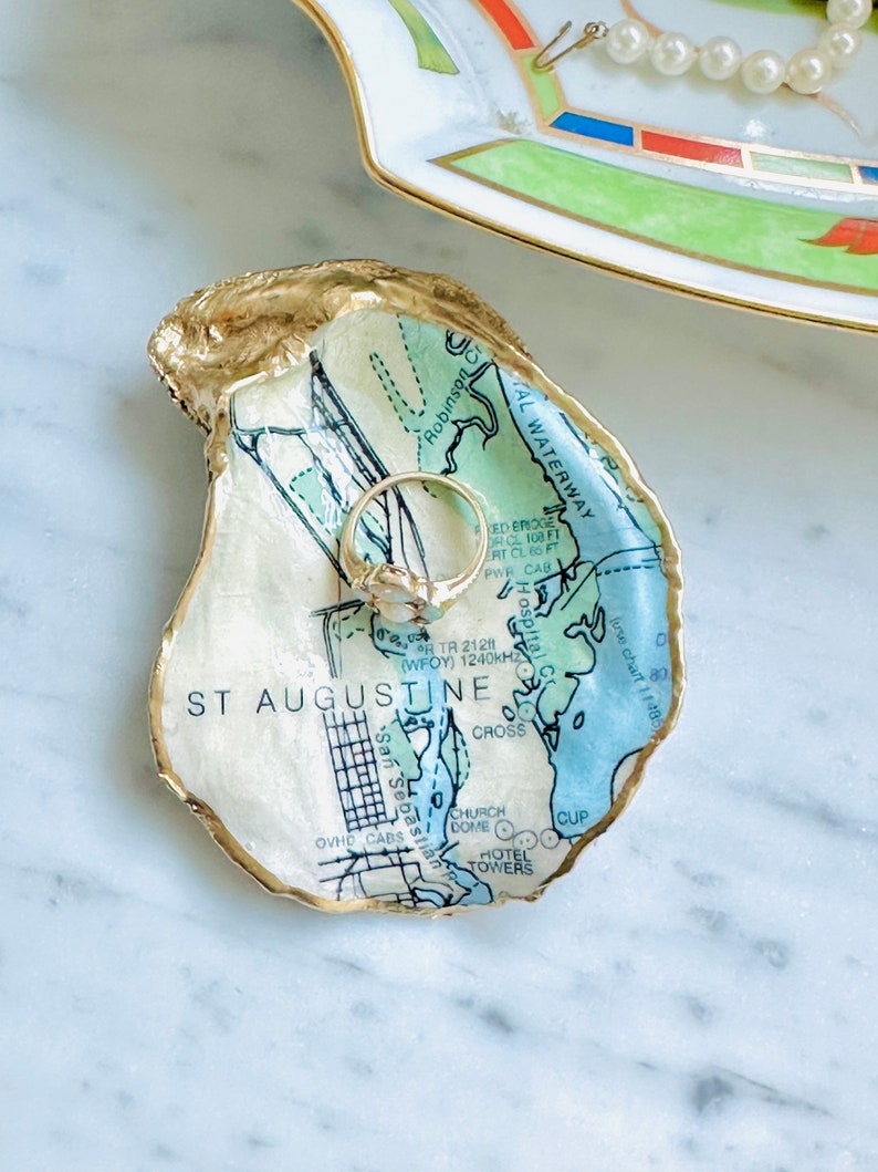 May include: An oyster shell ring dish featuring a map of St. Augustine, Florida. The shell has a gold-painted edge and holds a gold ring. The map design uses blue and green tones with black text. A decorative dish with a pearl necklace is in the background.
