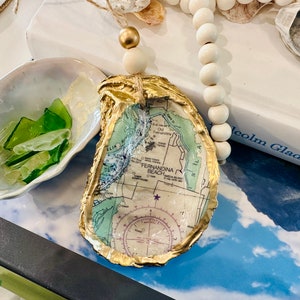 May include: A gold-painted oyster shell with a map of Fernandina Beach, Florida inside. The shell is hanging from a white beaded garland.