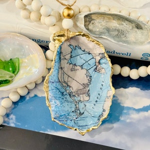 May include: An oyster shell decorated with a map of Tampa Bay, Florida, with a gold-painted edge. The shell is part of a coastal-themed display, with white wooden beads and other shells.