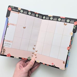 K&ouml;nnte beinhalten: Ge&ouml;ffneter Planer mit der Monats&uuml;bersicht f&uuml;r M&auml;rz. Der Planer hat ein Blumendesign mit Schmetterlingen. Auf der Oberseite der Seite steht "M&auml;rz" und es gibt einen Abschnitt f&uuml;r "Priorit&auml;ten". Der untere Teil der Seite hat einen Erinnerungsbereich.