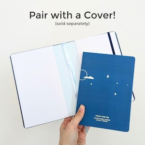 K&ouml;nnte beinhalten: Ein blaues Notizbuch mit Wolken- und Sterndesign wird neben einem offenen wei&szlig;en Notizbuch gehalten. Der Text "Pair with a Cover!" und "DESIGN YOUR LIFE" sind sichtbar. Das Notizbuch ist ein undatierter Wochenplaner.