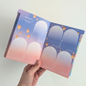 May include: Open planner with a gradient sky design, featuring the month of January. The pages display weekly layouts with arched sections for each day, adorned with floating lantern illustrations. The color scheme transitions from pink to blue.