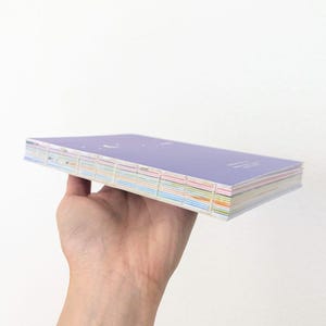 May include: A purple notebook with a stitched binding and multi-colored edges. The notebook is held in a hand against a white background. The cover has faint text.