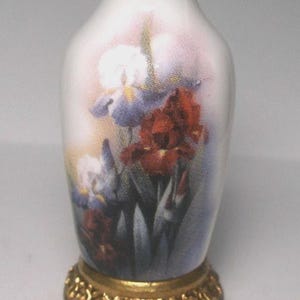 May include: A small white porcelain vase featuring a floral print of purple and deep red irises. The vase has a narrow neck with a gold-toned rim and is mounted on a circular, ornate gold-colored metal base with a textured, decorative edge.