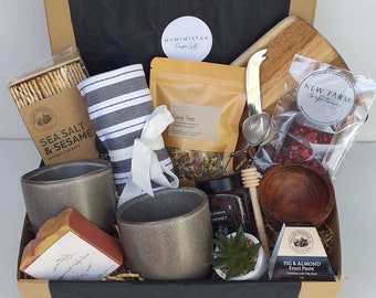 May include: A gift box filled with various gourmet food items, including artisan crackers, tea, honey, and fig and almond fruit paste. The box also includes a wooden cutting board, a tea infuser, and a small potted succulent plant.