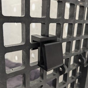 May include: A black 3D-printed plastic hook attachment secured onto a black grid panel. The hook features a flat, angled front surface and a U-shaped bracket that clips firmly over the vertical and horizontal bars of the square-patterned grid.