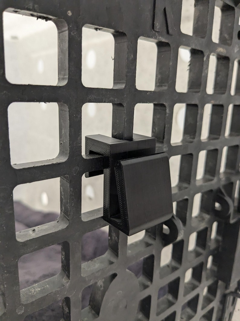 May include: A black 3D-printed plastic hook attachment secured onto a black grid panel. The hook features a flat, angled front surface and a U-shaped bracket that clips firmly over the vertical and horizontal bars of the square-patterned grid.