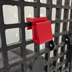 May include: A bright red plastic hook attached to a black grid panel. The hook features a flat top section that rests over the horizontal bar of the grid and a downward-angled front face, designed to hang items securely from the square-patterned frame.