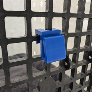 May include: A bright blue plastic hook designed to clip onto a black grid panel. The hook features a flat, angled front surface and a U-shaped bracket that secures firmly over the vertical and horizontal bars of the square-patterned grid structure.