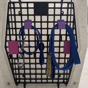 Puede incluir: Una puerta de perrera de pl&aacute;stico blanco moteado con una rejilla negra que incluye el texto Ruff Tough Kennels.com. Dos ganchos de pl&aacute;stico morado est&aacute;n montados en la rejilla, cada uno sosteniendo una correa azul. Dos soportes morados adicionales.