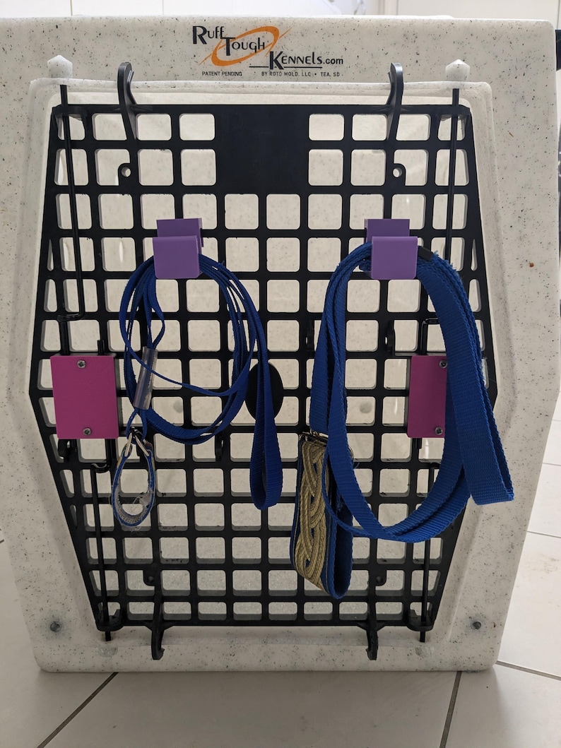 Puede incluir: Una puerta de perrera de pl&aacute;stico blanco moteado con una rejilla negra que incluye el texto Ruff Tough Kennels.com. Dos ganchos de pl&aacute;stico morado est&aacute;n montados en la rejilla, cada uno sosteniendo una correa azul. Dos soportes morados adicionales.