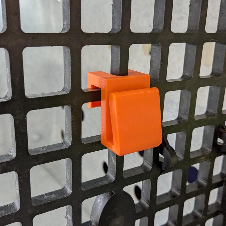 May include: A bright orange plastic hook designed to clip onto a dark gray grid panel. The hook features a U-shaped top section that fits over the vertical bar of the grid and a downward-facing hook component for hanging various items securely.