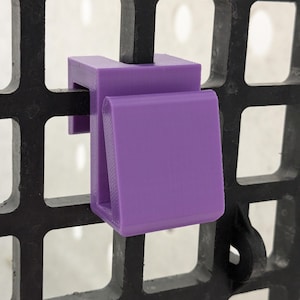 May include: A purple plastic hook designed to attach to a black grid wall panel. The hook features a U-shaped top section that fits over the grid wire and a downward-facing front tab for hanging items, constructed with a smooth, matte finish.