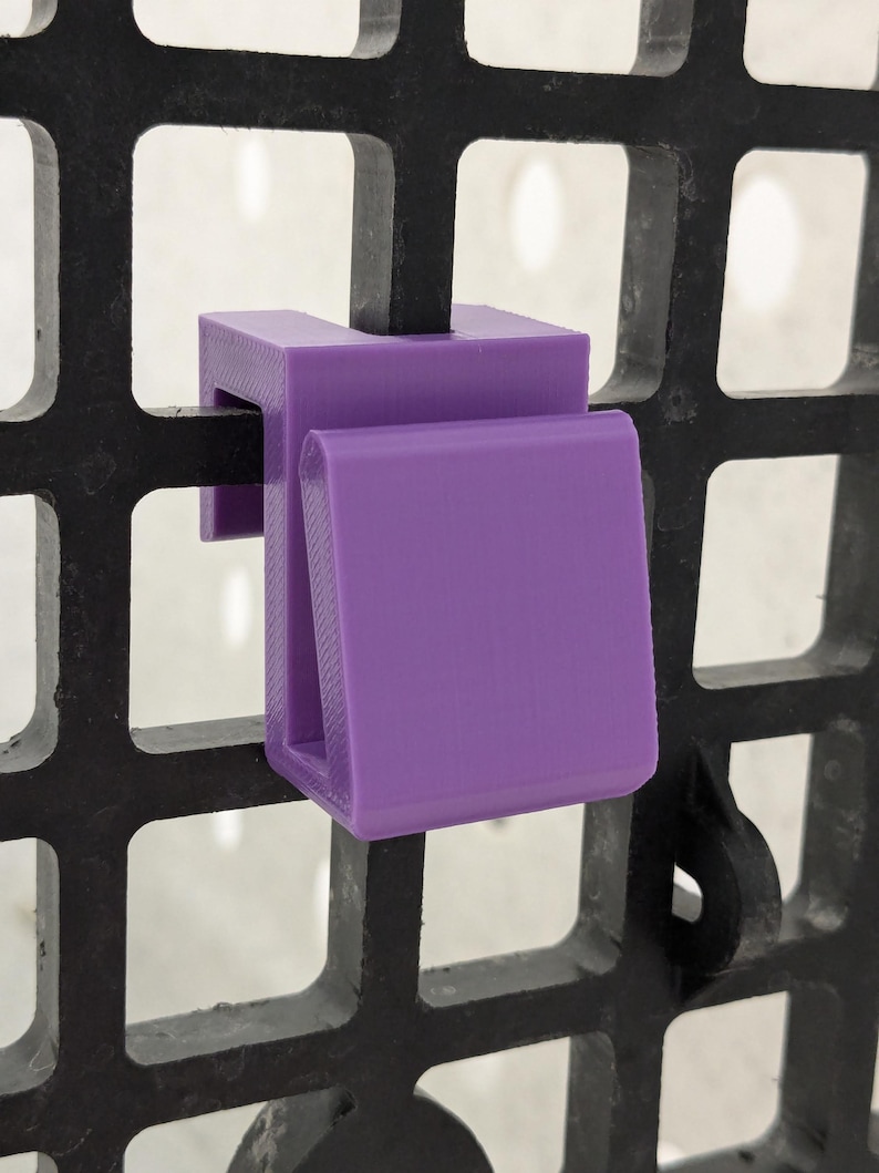 May include: A purple plastic hook designed to attach to a black grid wall panel. The hook features a U-shaped top section that fits over the grid wire and a downward-facing front tab for hanging items, constructed with a smooth, matte finish.