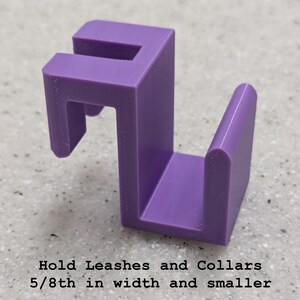 Puede incluir: Un gancho de pl&aacute;stico morado con un dise&ntilde;o de soporte para montaje, que incluye el texto: Hold Leashes and Collars 5/8th in width and smaller. El objeto tiene una estructura r&iacute;gida y angular con una ranura superior y una base en forma de U.