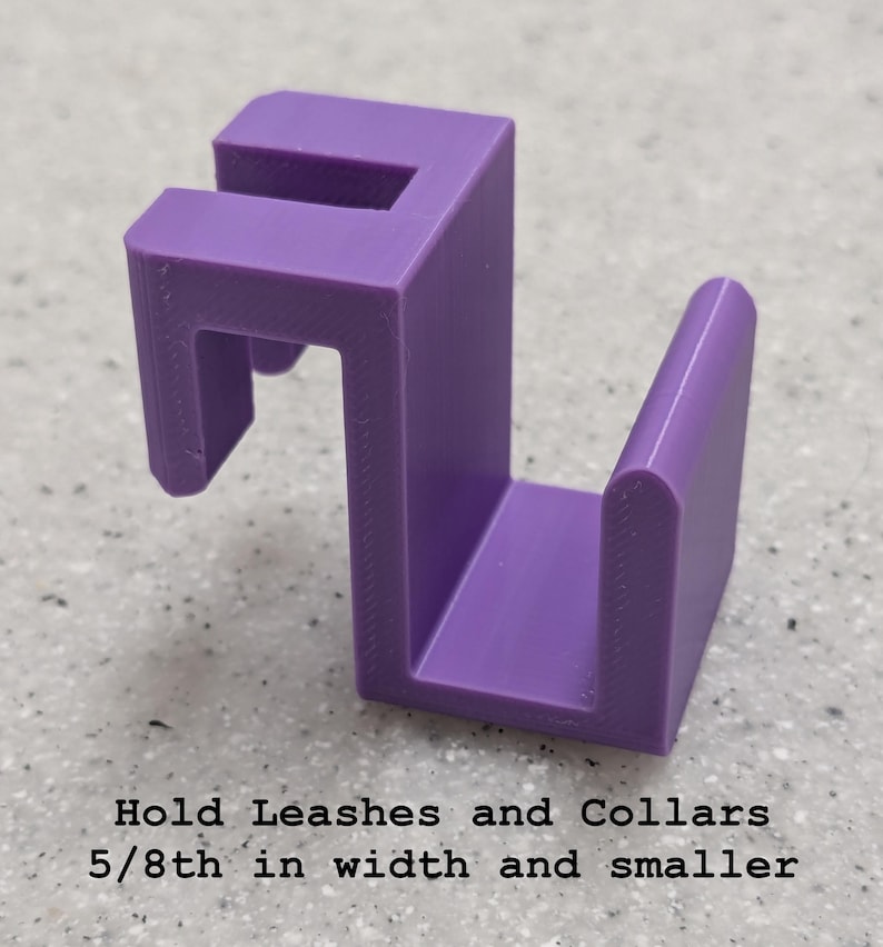 Puede incluir: Un gancho de pl&aacute;stico morado con un dise&ntilde;o de soporte para montaje, que incluye el texto: Hold Leashes and Collars 5/8th in width and smaller. El objeto tiene una estructura r&iacute;gida y angular con una ranura superior y una base en forma de U.