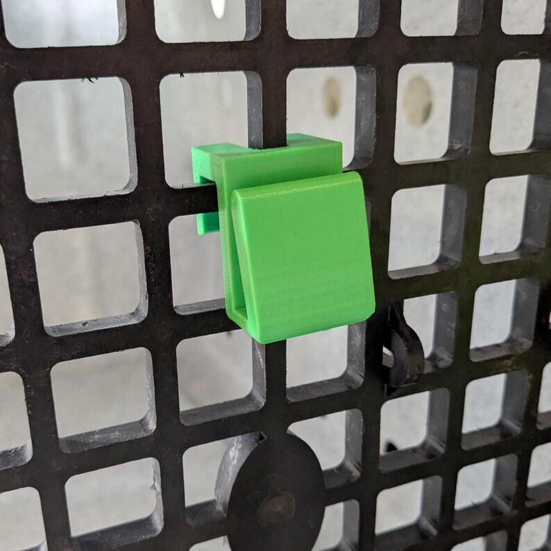 May include: A bright green plastic hook designed to attach to a black grid panel. The hook features a flat, rectangular front surface and a U-shaped clip mechanism that secures firmly onto the square openings of the dark grid structure.