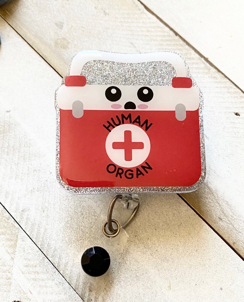 K&ouml;nnte beinhalten: Ein silberner Glitzer-Badgehalter mit einem roten Erste-Hilfe-Kasten-Design. Der Kasten hat ein wei&szlig;es Kreuz und die Worte "HUMAN ORGAN" darauf. Der Kasten hat ein s&uuml;&szlig;es Comic-Gesicht mit gro&szlig;en Augen und einem &uuml;berraschten Ausdruck.
