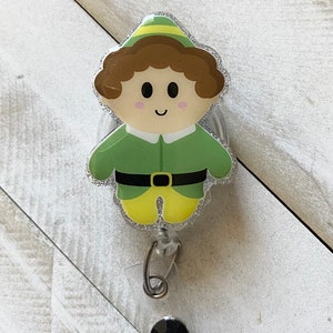 May include: A green and yellow glitter badge holder featuring a cartoon elf with brown hair and a green hat. The elf is smiling and has a black belt.