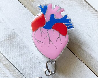 May include: A pink anatomical heart with red and blue details, with glitter accents, attached to a clear plastic retractable badge holder.