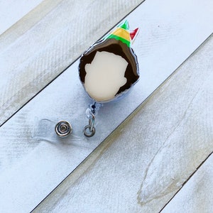May include: A clear plastic badge holder with a cartoon elf design. The elf is wearing a green and red hat and has brown hair. The badge holder has a clear plastic strap and a metal clip.
