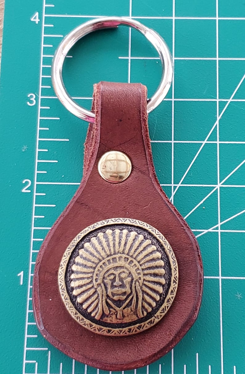 May include: A brown leather keyring with a silver key ring. The keyring features a brass medallion with an image of a Native American chief. A brass rivet secures the leather loop to the key ring.