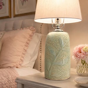 Puede incluir: Una lámpara de mesa con base cilíndrica de cerámica en tono verde pálido, con una superficie texturizada con patrones de hojas en relieve. La lámpara tiene una pantalla de tambor de tela color blanco roto y un cuello de metal plateado.