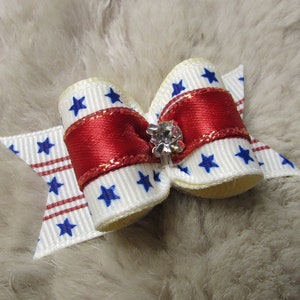 May include: A red, white, and blue star-patterned bow with a sparkly red center and a clear rhinestone in the center. The bow is on a white furry surface.