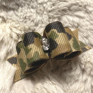 May include: A hair bow made of ribbed fabric featuring a camouflage pattern in shades of olive green, brown, and black. The center of the bow is cinched with a small, silver-toned decorative element encrusted with clear, sparkling rhinestones.