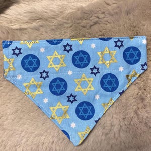 May include: A light blue dog bandana with a repeating pattern of gold and blue Star of David symbols, blue circles with the Star of David, and white stars. The bandana is triangular in shape.
