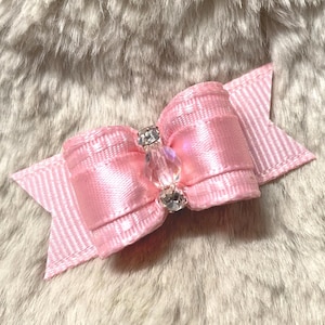 May include: A pink satin bow with a crystal and rhinestone center. The bow has a layered design with a striped ribbon base. The bow is set against a soft, textured background.