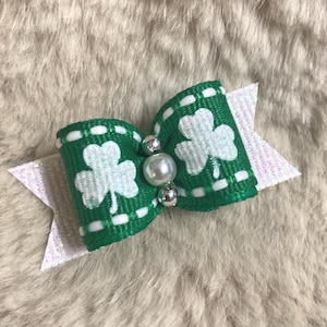 May include: A green and white bow featuring a shamrock design, perfect for St. Patrick's Day. The bow has a white glittery tail and is adorned with a pearl and silver beads. The bow is approximately 3 inches wide.