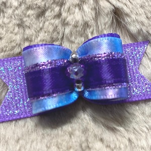 May include: A purple and blue satin bow with a glittery purple accent. The bow features a central band of deep purple, adorned with a small, clear gem and silver beads. The bow's ends are shaped like a star.