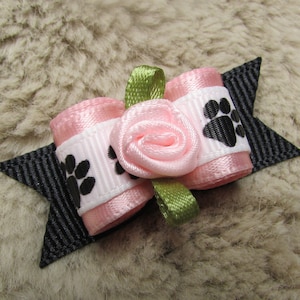 May include: A pink and white bow with black paw prints and a pink rose in the center. The bow is attached to a black clip.