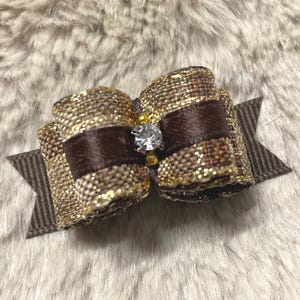 May include: A decorative hair bow constructed from brown and gold metallic woven fabric, featuring a central dark brown satin ribbon band accented with a clear faceted rhinestone and two small gold beads at the center of the bow structure.