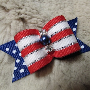 May include: A patriotic-themed bow featuring red and white striped ribbon with silver trim, a navy blue pearl, and a blue star-shaped base with white polka dots. This decorative bow is suitable for pets.